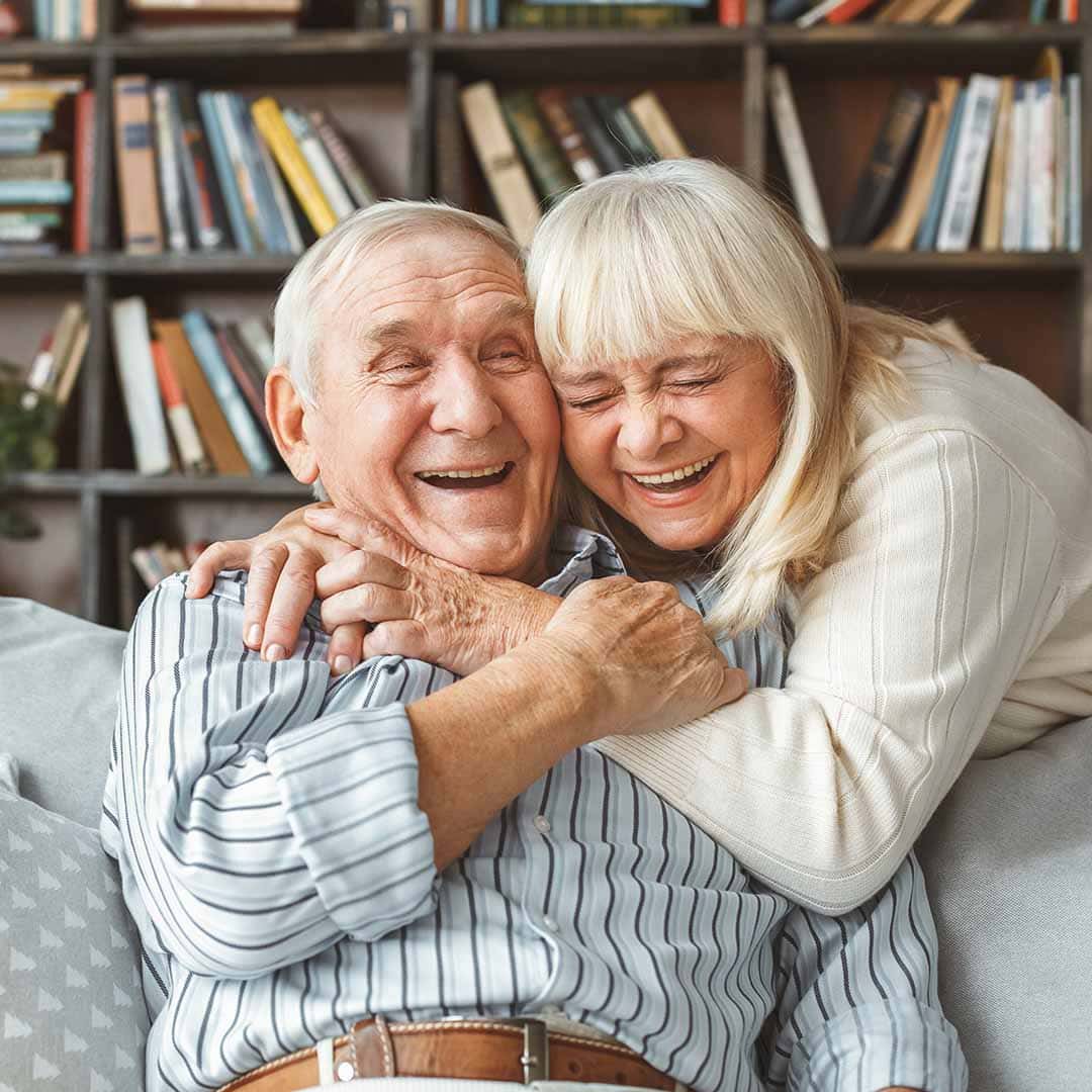 Portrait Photo of Senior Couple Hugging and Laughing while sitting on a Couch at Stellar Living Communities